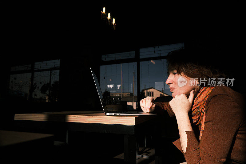 一个开朗的成熟女人坐在昏暗的咖啡馆里，用笔记本电脑和别人聊天