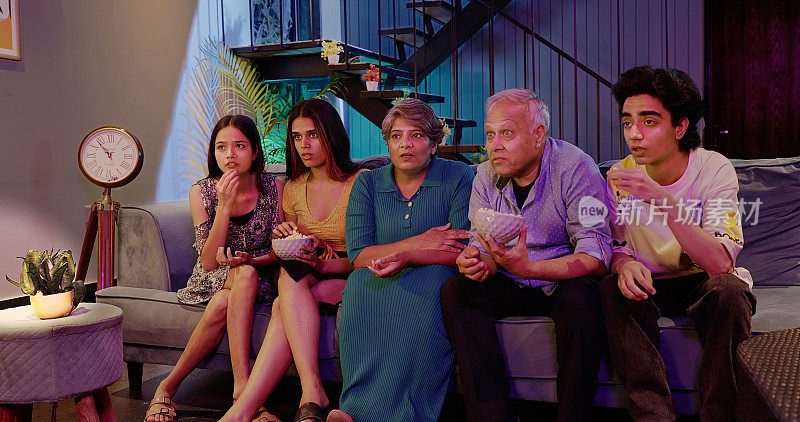 印度印度教延伸幸福的家庭坐在沙发上看恐怖惊悚电视害怕的脸祖父母吃零食年轻的成年男孩十几岁的女孩吃零食拿爆米花碗人们一起深夜在家里看电影