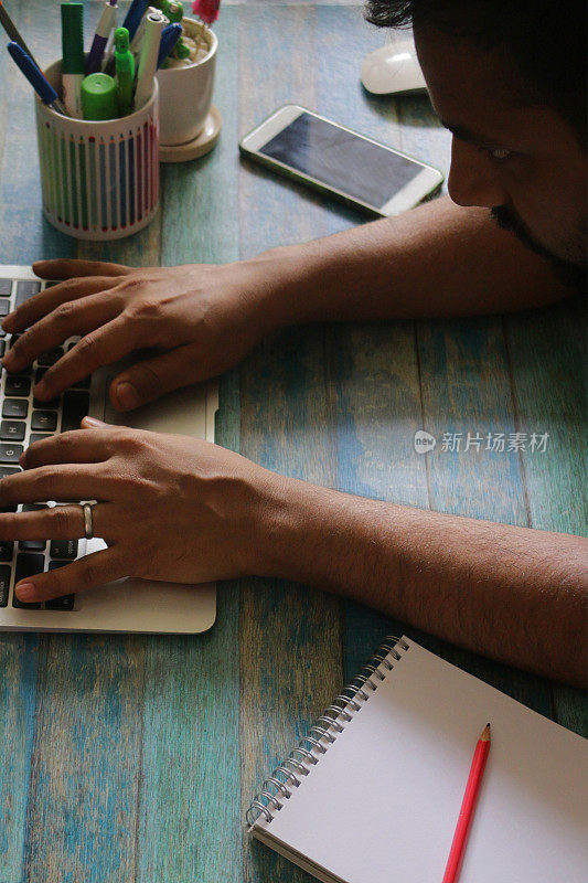 图片中，一名亚洲男性在办公室工作站的笔记本电脑上打字，周围环绕着无线鼠标、笔筒、盆栽仙人掌、智能手机、笔记本和铅笔，蓝色的木纹背景，聚焦前景