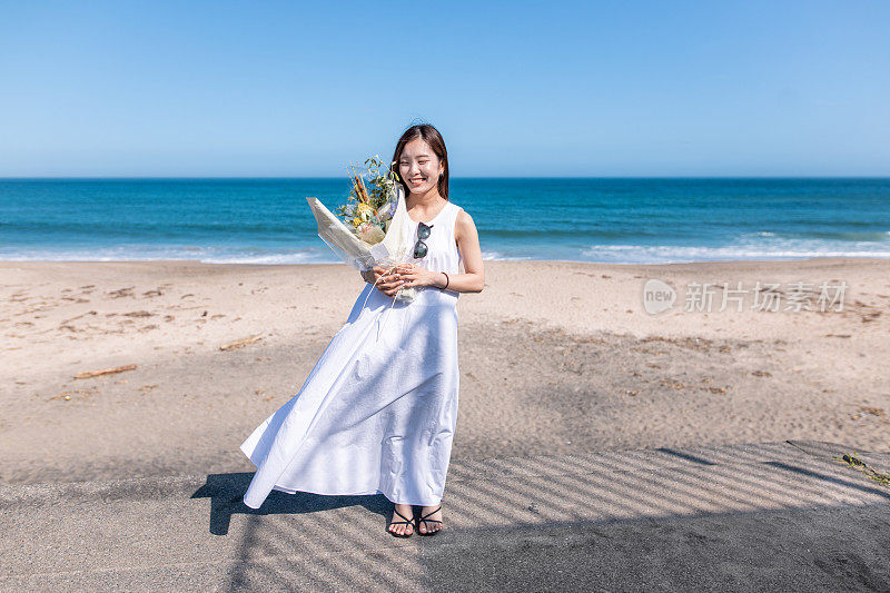 年轻女子的肖像站在夏天的海滩上，拿着花束