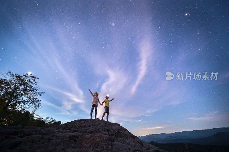 一起去摘星星。宽阔的镜头一对相爱的游客站在一个高岩石在晚上，看着宏伟的星空。灵感和灵性。心理健康。