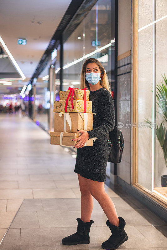 新冠肺炎疫情期间，年轻美丽的女孩在商场享受圣诞购物。她戴着防护口罩，以防止冠状病毒感染。