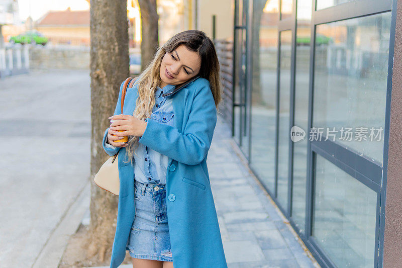 年轻漂亮的女人拿着一杯咖啡走在街上。迷人的年轻女性在时尚的衣服拿着一杯热饮