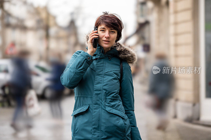 年轻女子穿着蓝色雨衣在户外使用手机在城市街道人行道上