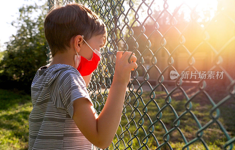 这个小男孩心情很不好，他戴着一个保护面罩，透过学校的栅栏看着，一边摇晃着