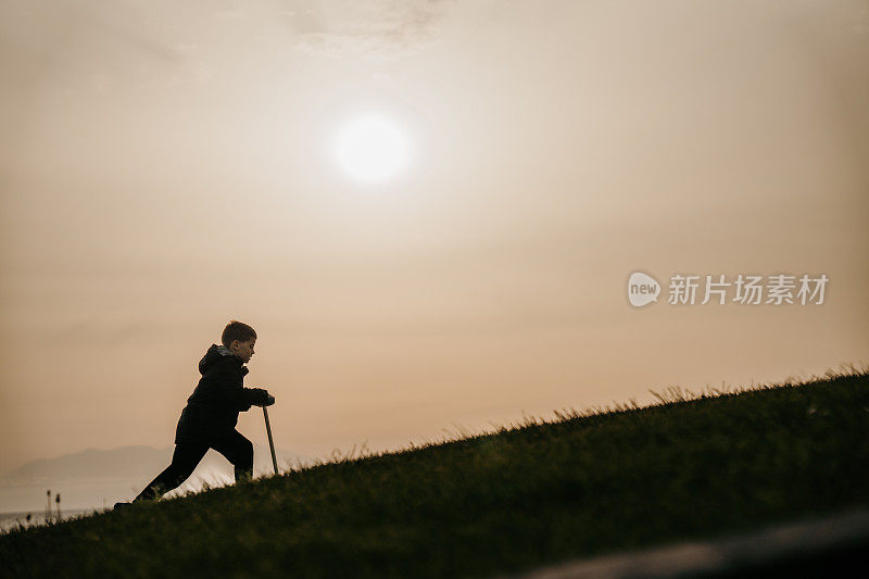 日落时分，小男孩推着他的踏板车走在海边的小山上