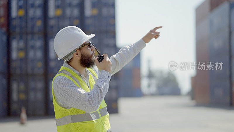 工程师或工头穿着安全帽安全背心，通过对讲机通话，控制集装箱堆场的货物装载和装运过程，以便将货物运送到支持物流的货船上。