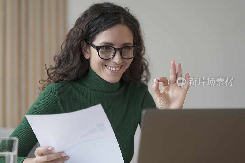 微笑的意大利女员工手持财务报告，并在视频通话中显示okay手势