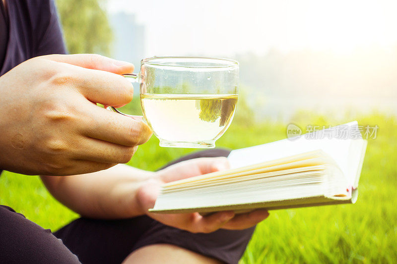 女人坐在草地上，手里拿着茶和书