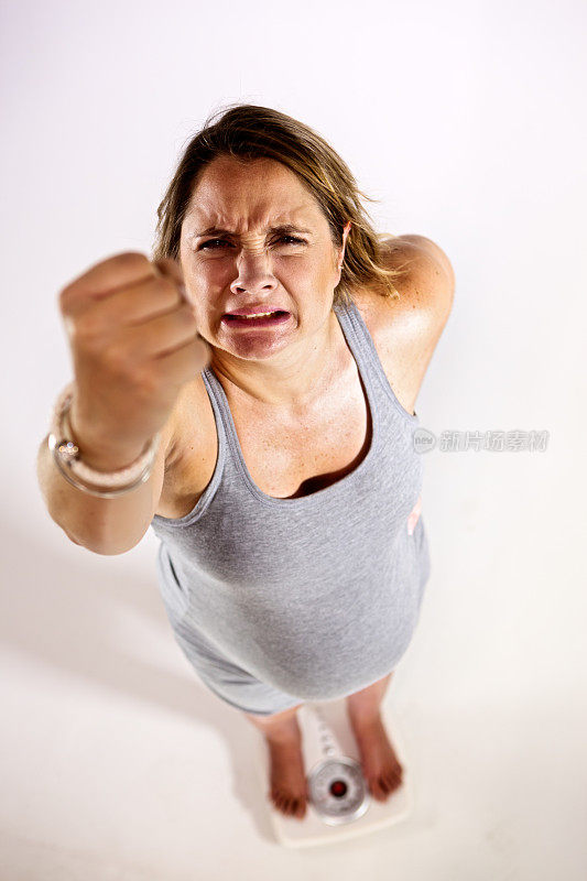 磅秤上愤怒的孕妇挥舞着拳头。高角度。