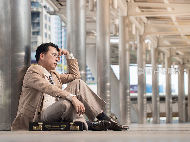 穿着西装的亚洲商人带着黑色公文包坐在办公室外，焦虑而紧张。失业或经济概念。