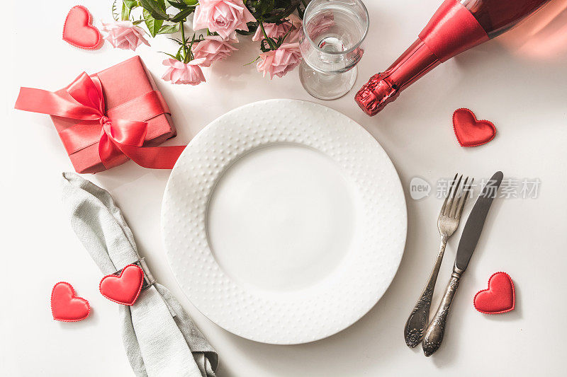 情人节晚餐与桌子的位置设置红色礼物，玻璃为香槟，一瓶香槟，粉红玫瑰，心形饰品与银器在白色的背景。前视图。情人节卡片。