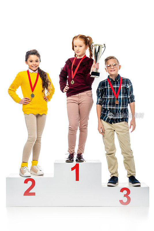 快乐的孩子与奖章和奖杯站在胜利者基座上，微笑着看着相机孤立在白色