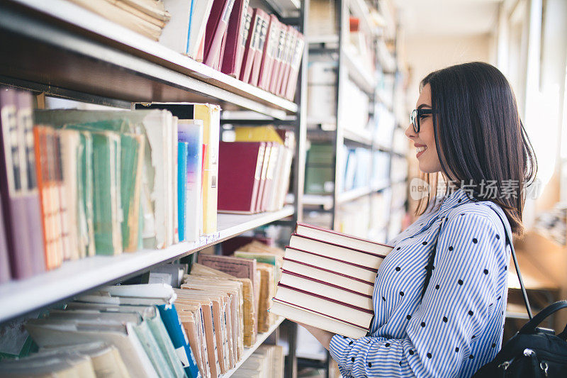 年轻的学生拿着书和背包站在大学图书馆的书架旁