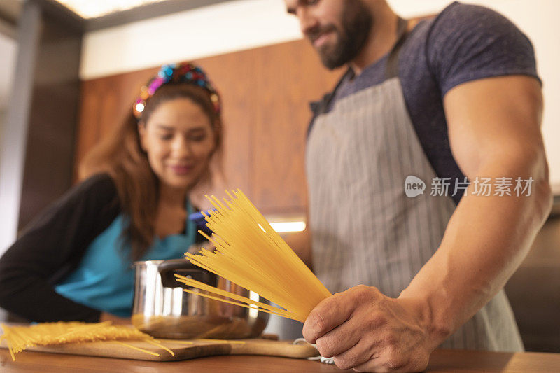 一对夫妇穿着围裙的男人和女人在厨房里一起准备食物，做意大利面。看如何用智能手机做饭的视频。