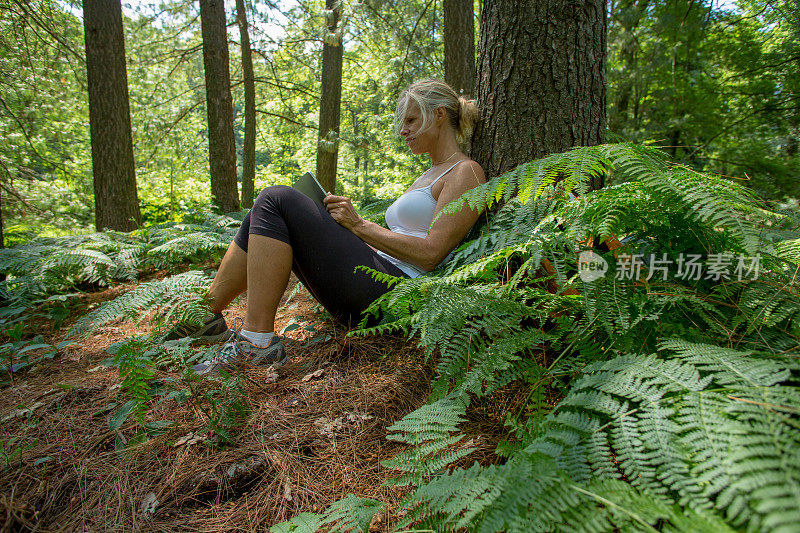 一个成熟的女人在森林里靠在树上，用着平板电脑