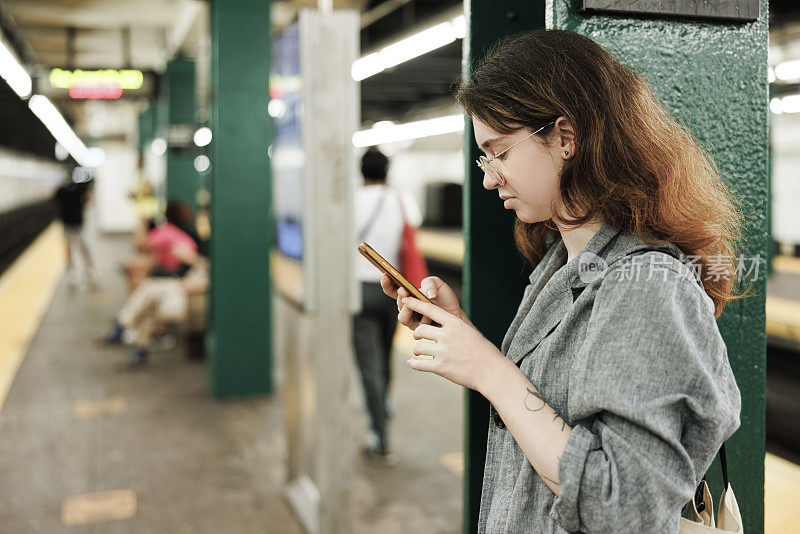 地铁高峰时段。一名戴眼镜的卷发女子在地铁站台上用智能手机上网。