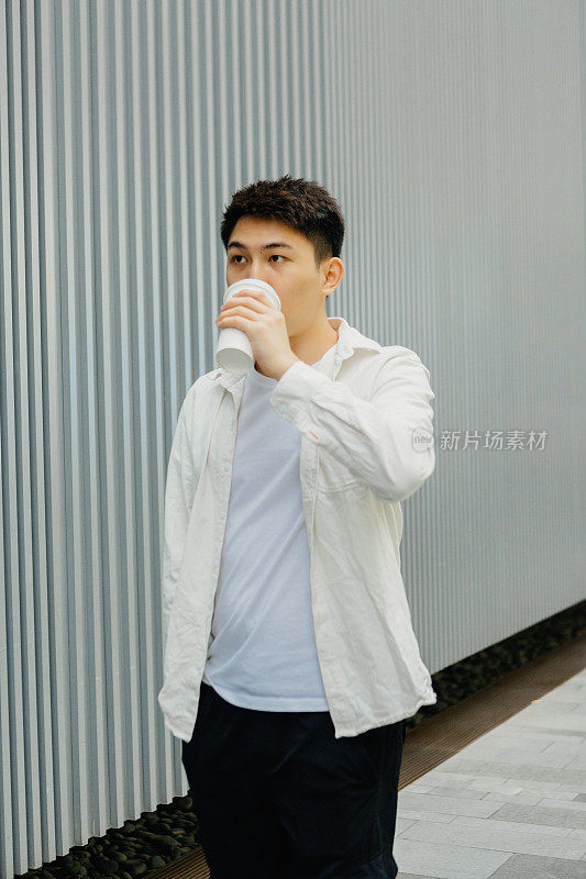 年轻的亚洲男性商人正在喝着咖啡，走在灰色的户外墙壁前——下班后喝一杯咖啡，促进健康，工作生活平衡