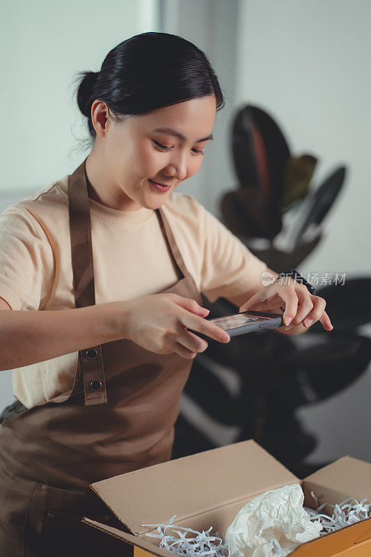 亚洲女卖家在打包货物准备发货前用智能手机拍照。