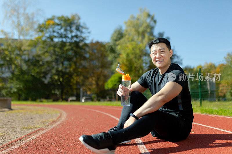 年轻的亚洲运动员在慢跑、运动后坐在跑步机上休息。他拿着瓶子，喝水，恢复精神，解渴。微笑着看着镜头