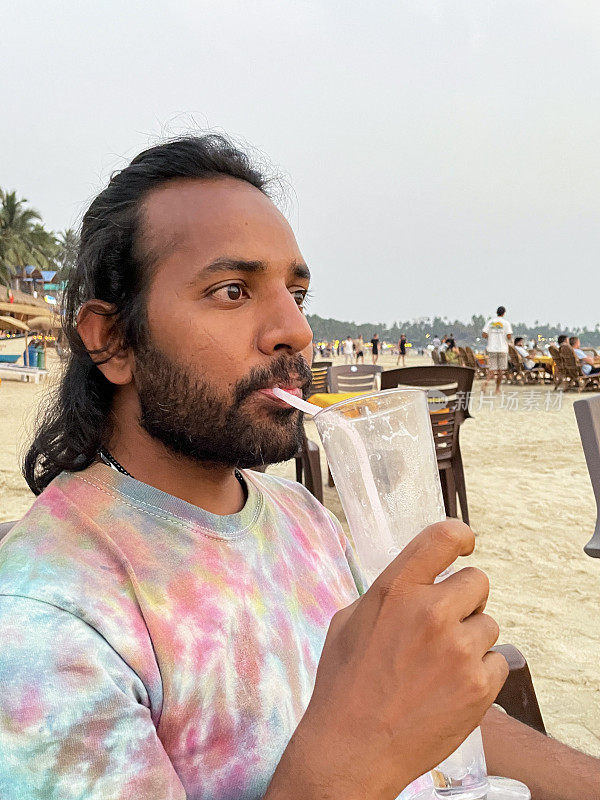 海滩餐厅酒吧场景特写，印度男子坐在露天用餐区的餐桌旁，用弯曲的吸管从玻璃杯中啜饮鸡尾酒，聚焦于前景