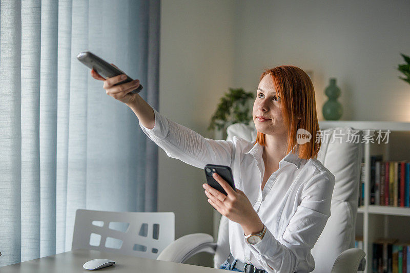 一位女企业家手持手机和遥控器在办公室调节空调，运用科技理念