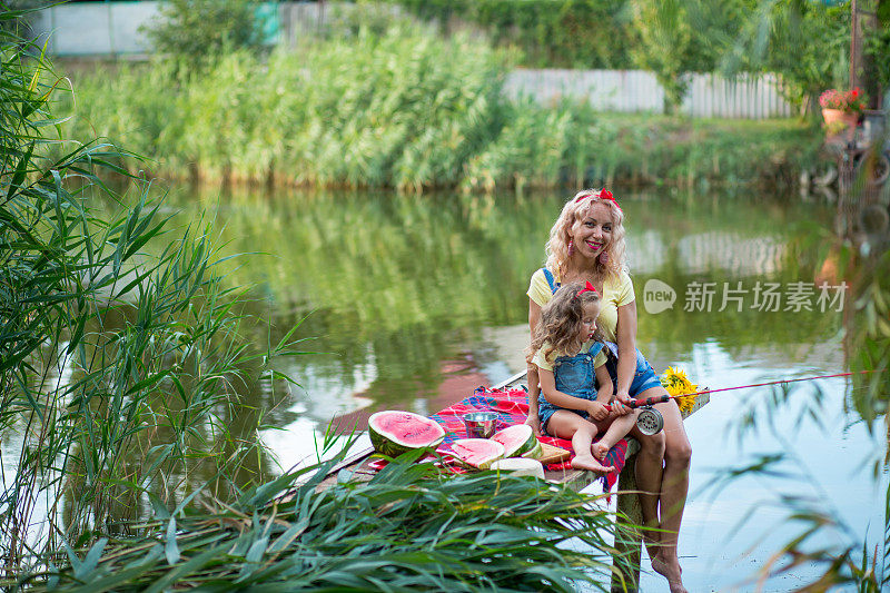 金发碧眼的母亲和三岁的女儿穿着牛仔短裤，系着红色蝴蝶结的腰带，在河上的码头上吃西瓜和鱼