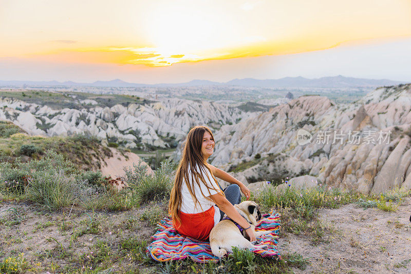 快乐的女人和她的狗在土耳其卡帕多西亚风景秀丽的山谷里欣赏日落