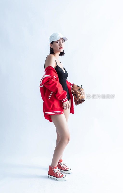 一位戴着棒球帽、穿着夹克、泳衣和运动鞋的年轻女子拿着棒球手套。