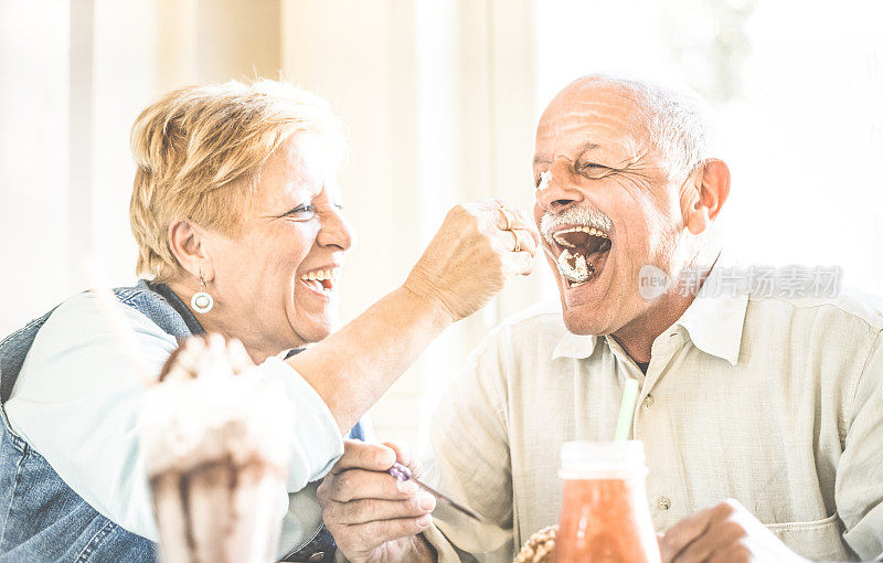 快乐的退休老夫妻在享受生物冰淇淋杯-快乐的老人生活理念-妻子喂丈夫和在常青假期在酒吧咖啡厅餐厅的乐趣-复古滤镜