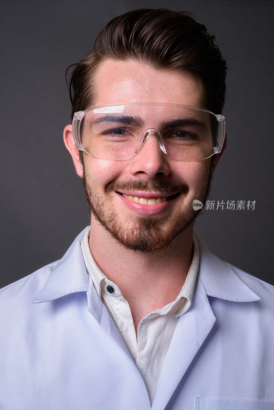 年轻的胡子英俊的男子医生戴着保护眼镜对抗灰色的背景