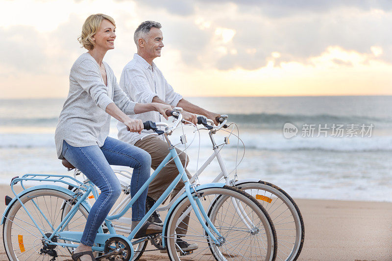 成熟的情侣在日落或日出时在海滩上骑自行车。