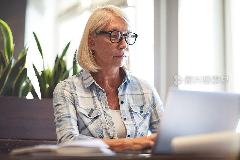 严肃、专注、美丽、成熟的女人戴着眼镜坐在桌子前，一边在小咖啡馆里写报告，一边在笔记本电脑上打字