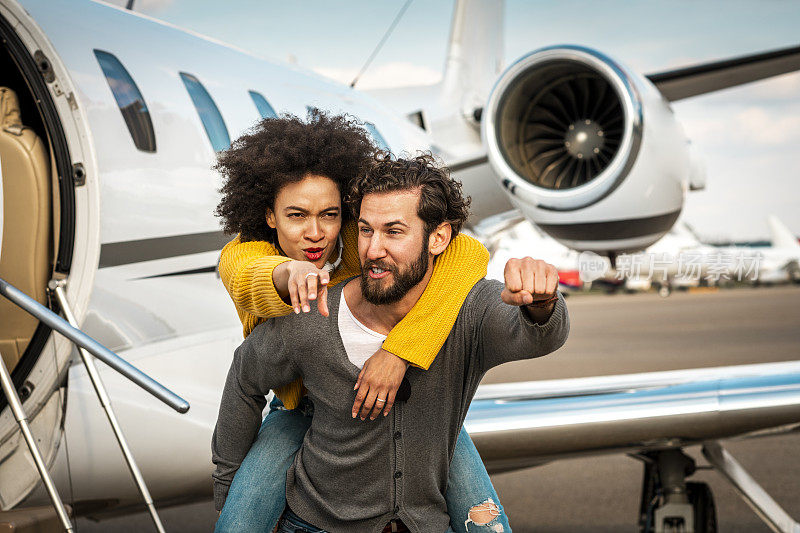 年轻的成功女性，她的伴侣背着她坐在停在机场跑道上的私人飞机旁边