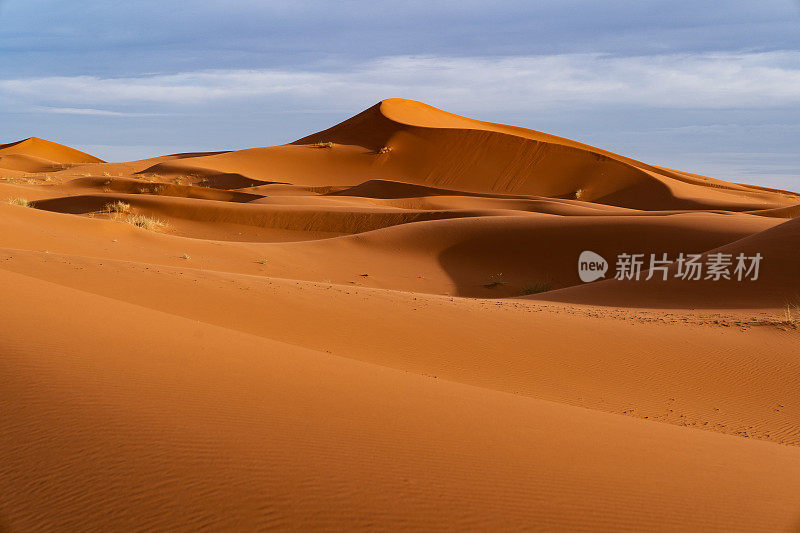 巨大的橙色沙状沙丘和多云的天空在Merzouga沙漠，摩洛哥。