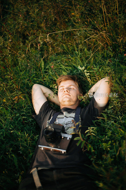 时髦的潮人与电影相机躺在绿色的草地上，放松在阳光灿烂的草地在大气的日落。帅哥在大自然中休息，在山中享受夜晚