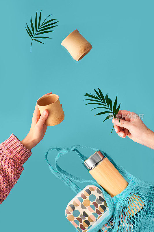 环保保温竹瓶零废茶。两只手拿着杯和棕榈叶，网袋和午餐盒和茶瓶飞。零废物概念悬浮在青色背景。