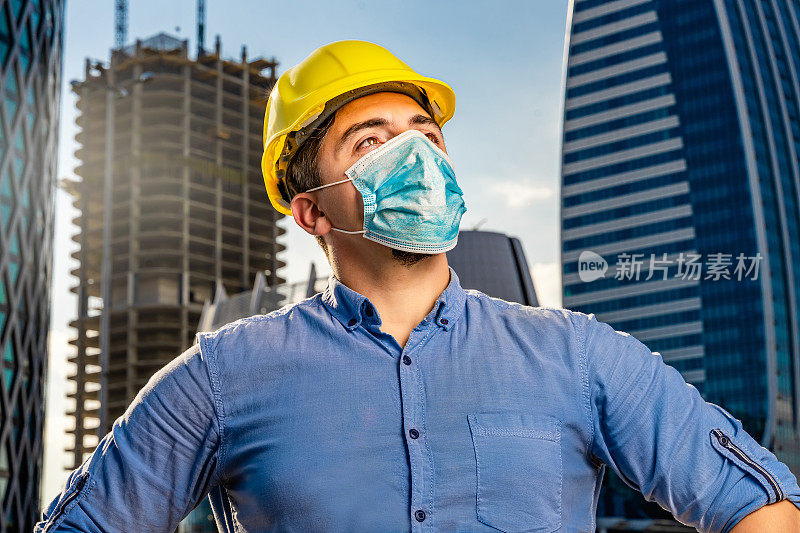 年轻工程师戴着黄色安全帽和N95防护口罩，站在现代化的商业建筑和建筑工地前