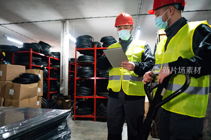 带防护面罩的男性仓库工人，用数码板和托盘千斤顶对仓库内健身用成品橡胶垫进行分类
