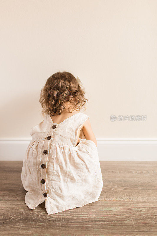 2019冠状病毒病(COVID-19)期间，两岁蹒跚学步的小女孩蹲在地板上，身穿奶油色亚麻裙，背部有纽扣和蝴蝶结