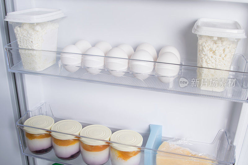 在白色冰箱的门上，架子上放着鸡蛋、奶酪、酸奶和白软干酪