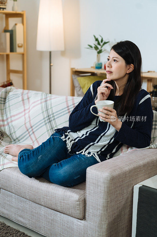 垂直拍摄的女人懒洋洋地躺在沙发上，手里拿着热饮料。全长漂亮的亚洲女士正在思考与手放在下巴，而喝咖啡。真正的时刻