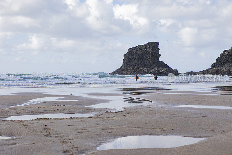 两个带着冲浪板的人走在安静的海滩上，远处有岩石，天空多云