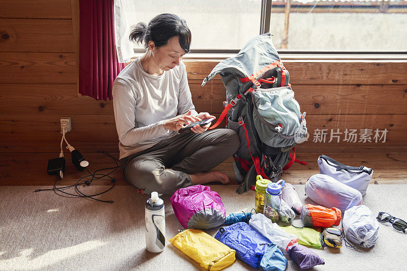 一名亚洲女子在山间小屋收拾行李时，用智能手机查看明天的天气和登山路线。