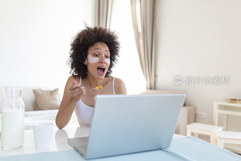 年轻迷人的女人坐在室内的桌子旁，手提电脑上放着玉米片。看着笔记本电脑和她的朋友通过视频通话。