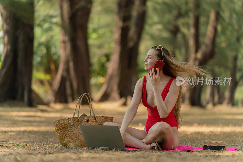 美丽的白人女性性感的女人在红色泳装比基尼与太阳镜在她的电脑笔记本电脑上工作，并在她的手机上聊天，而坐在海边的树林放松