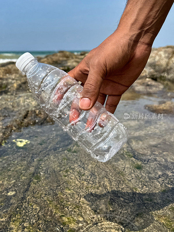 特写图片，在退潮时，白色盖子的空透明塑料水瓶被不认识的人捡起，海岸上的海洋垃圾和污染，乱扔的沙子，肮脏的海滩，重点在前景