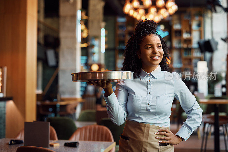 快乐的黑人女服务员端着托盘在咖啡馆里看向别处。