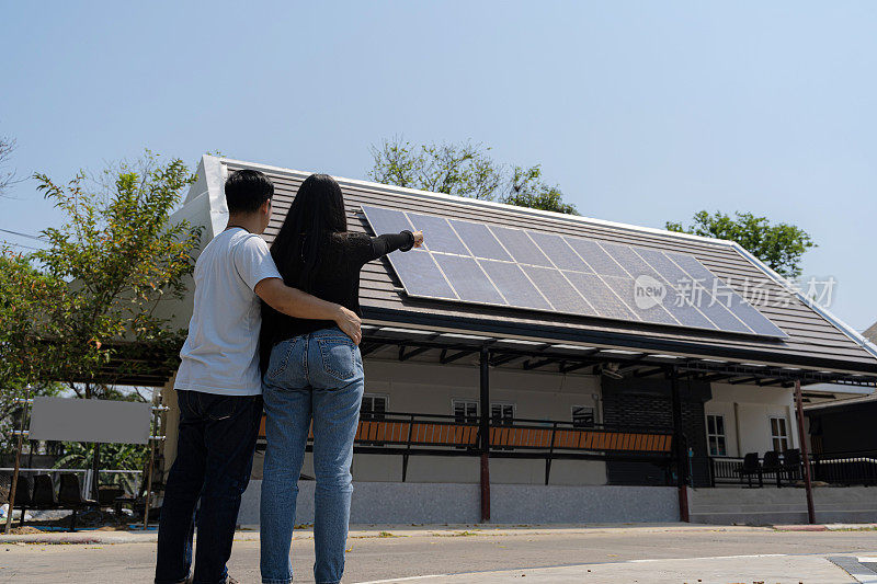 家庭使用可再生能源系统与太阳能电池板。一对年轻夫妇站在他们的新房子前的后景