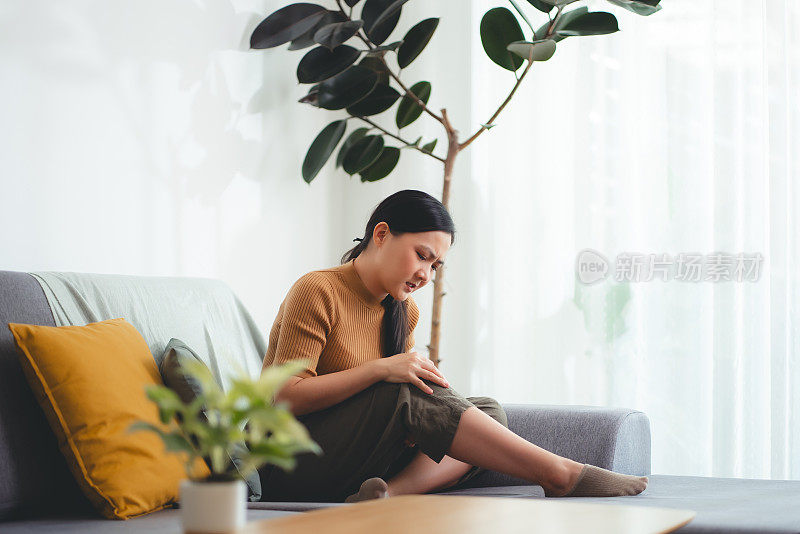 一位亚洲妇女坐在家里客厅的沙发上忍受着膝盖疼痛。